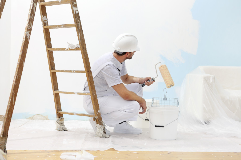 Man in overalls repainting a blue wall white with a roller brush